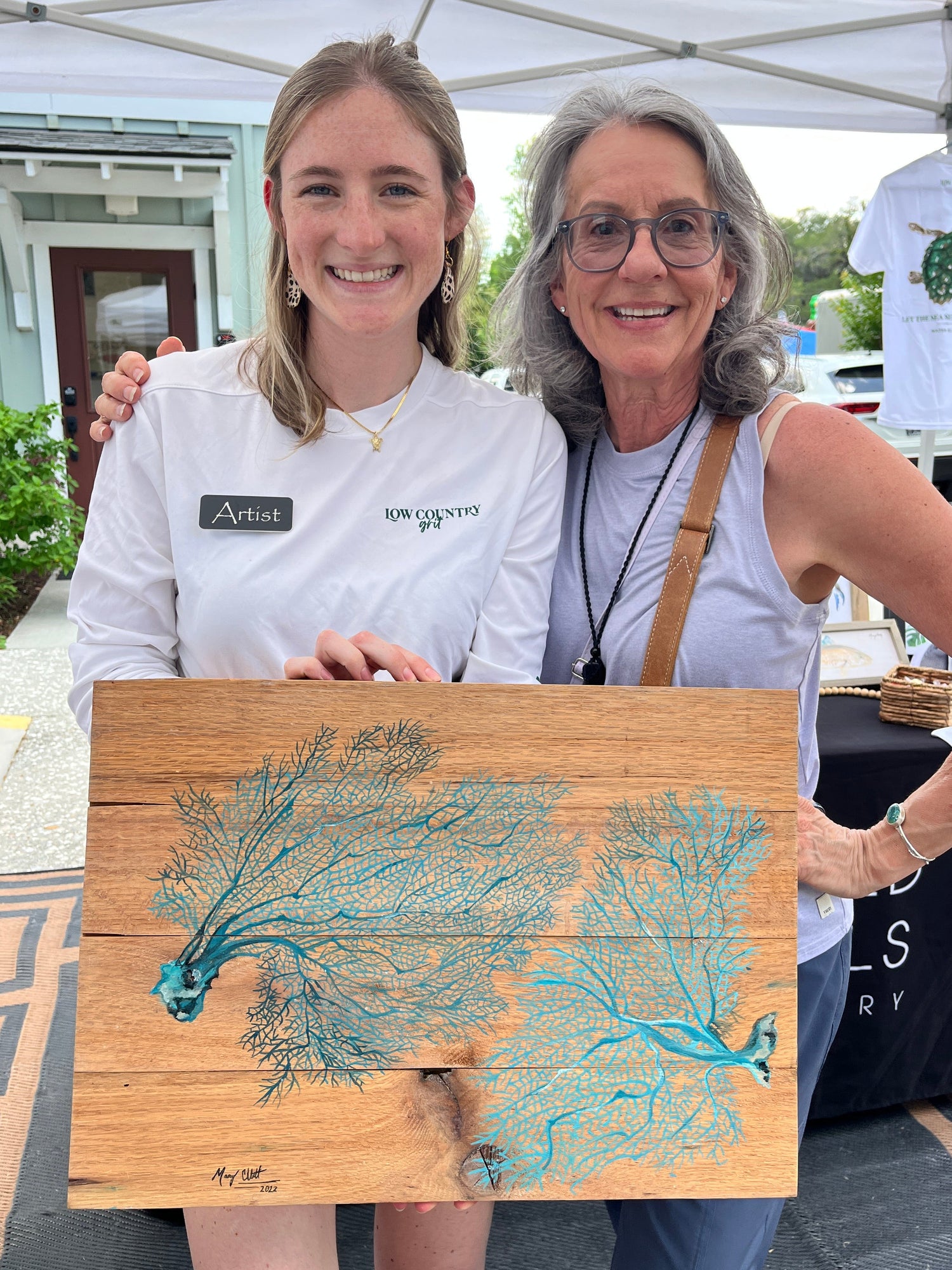 Two women holding a wooden board with a blue and green design outdoors.